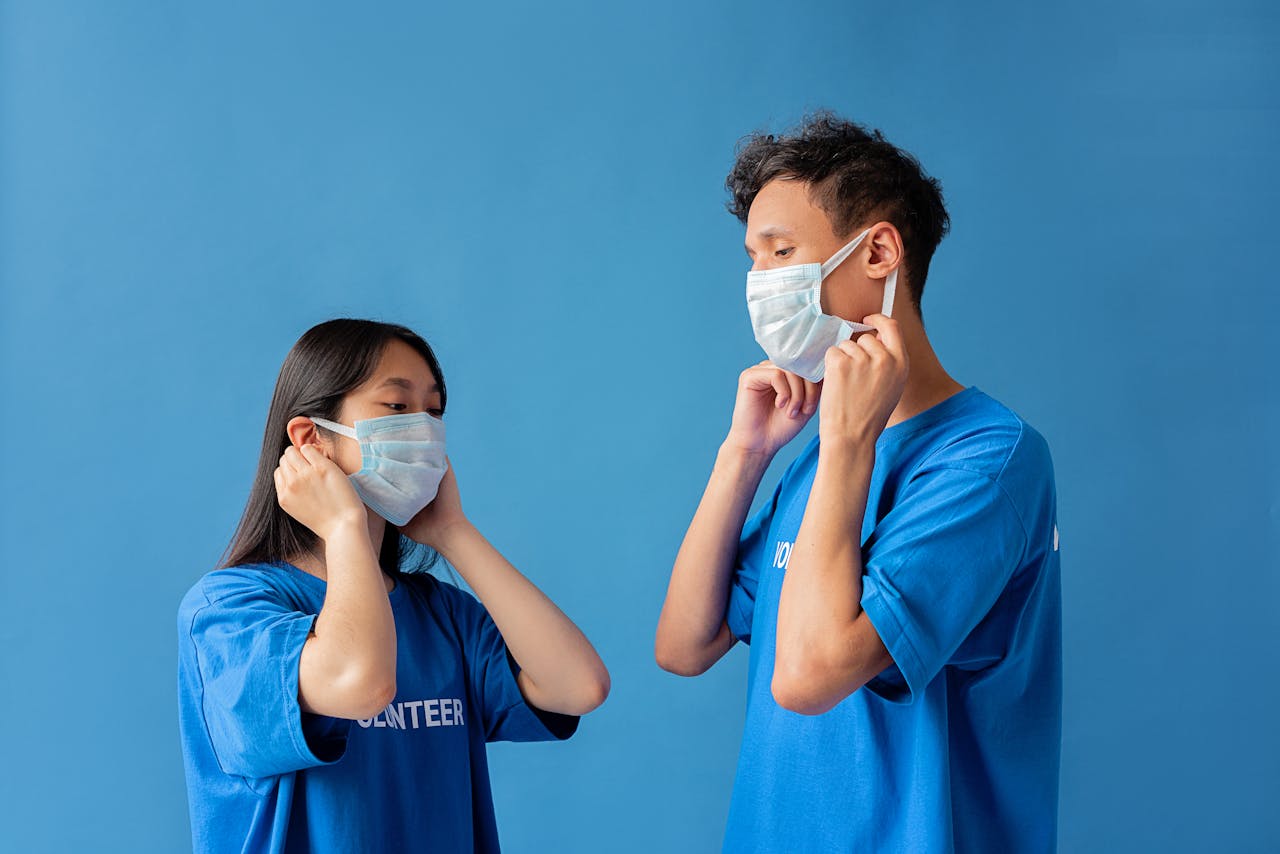 Two young volunteers wearing blue shirts and face masks against a blue background, preparing to help.